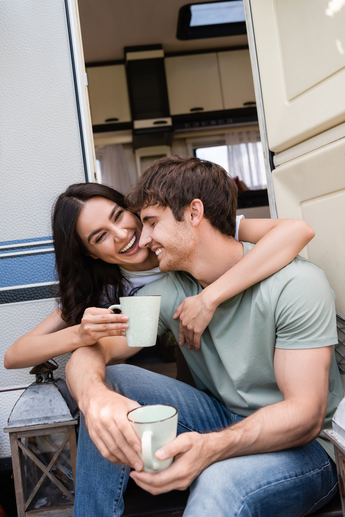Positive young woman hugging boyfriend with cup near door of camper van  - Photo, Image