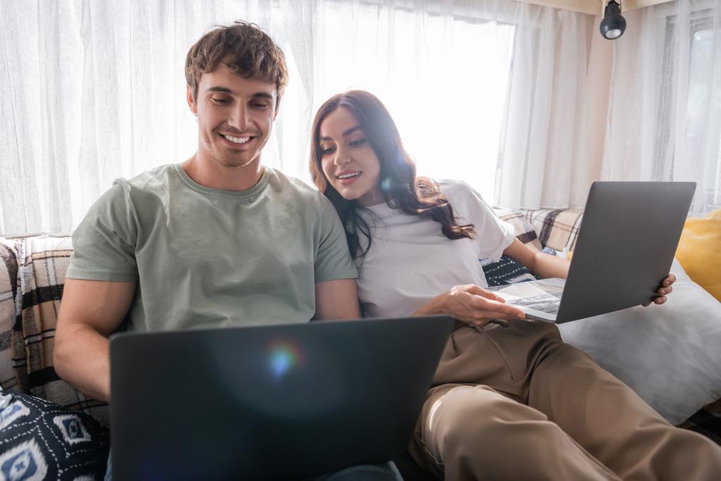 Positive woman looking at laptop near blurred boyfriend on bed in camper van  - Foto, afbeelding