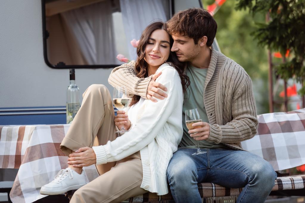 Brunette man in cardigan hugging girlfriend with wine on terrace of camper van  - Zdjęcie, obraz