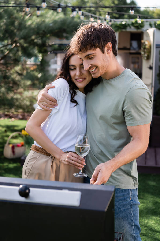 Smiling man hugging girlfriend with wine near blurred grill outdoors  - Fotoğraf, Görsel