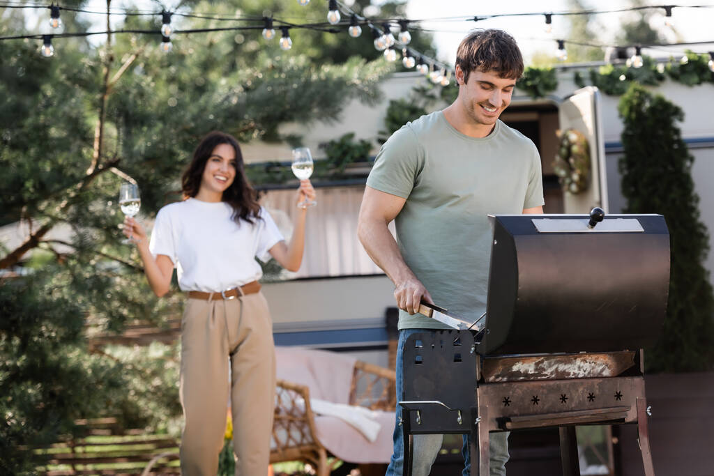 Smiling man cooking on grill near blurred girlfriend with wine and camper van  - Foto, imagen