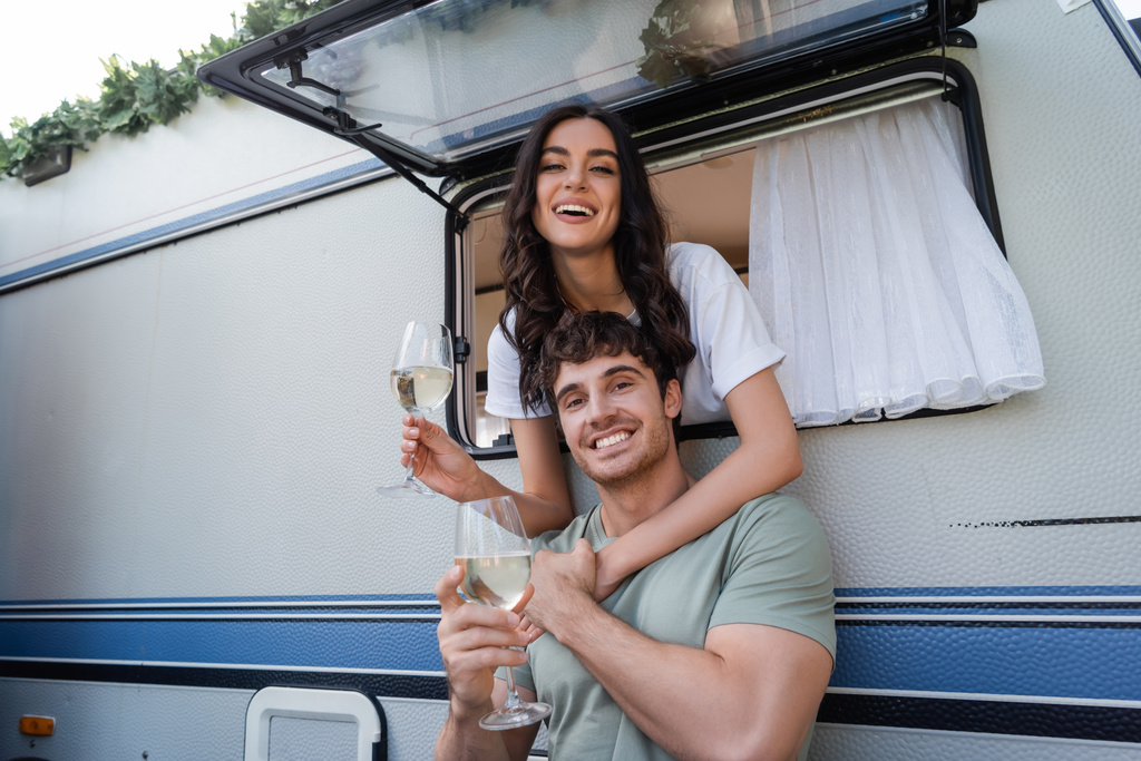 Smiling brunette woman hugging boyfriend with glass of wine near camper van  - Foto, afbeelding