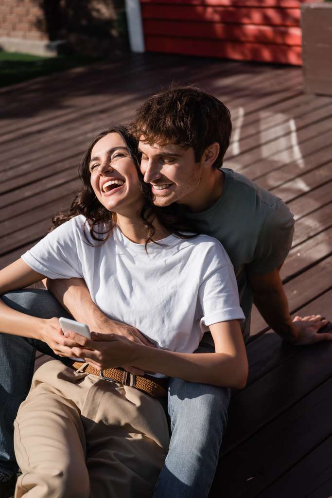 Cheerful man hugging brunette girlfriend with smartphone on terrace  - Valokuva, kuva