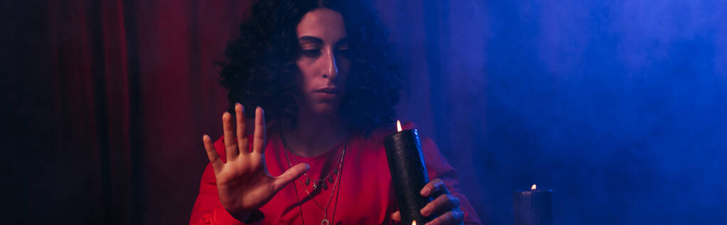 young oracle holding burning candle during spiritual session in darkness with blue smoke, banner  - Fotoğraf, Görsel