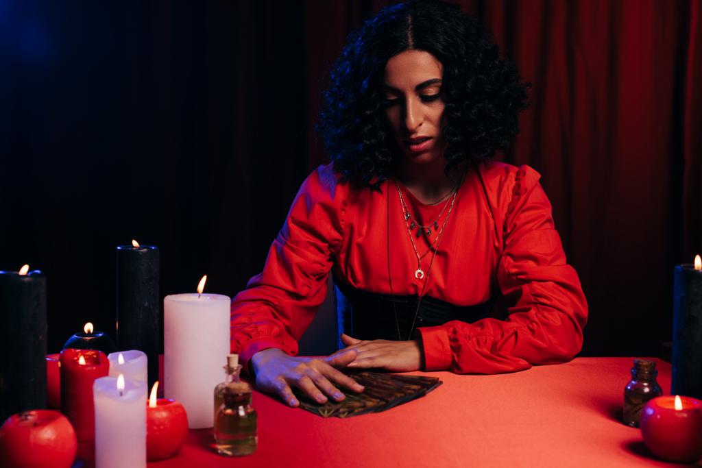 молода брюнетка предиктор біля карт таро і палаючі свічки на червоному столі і темному фоні
 - Фото, зображення