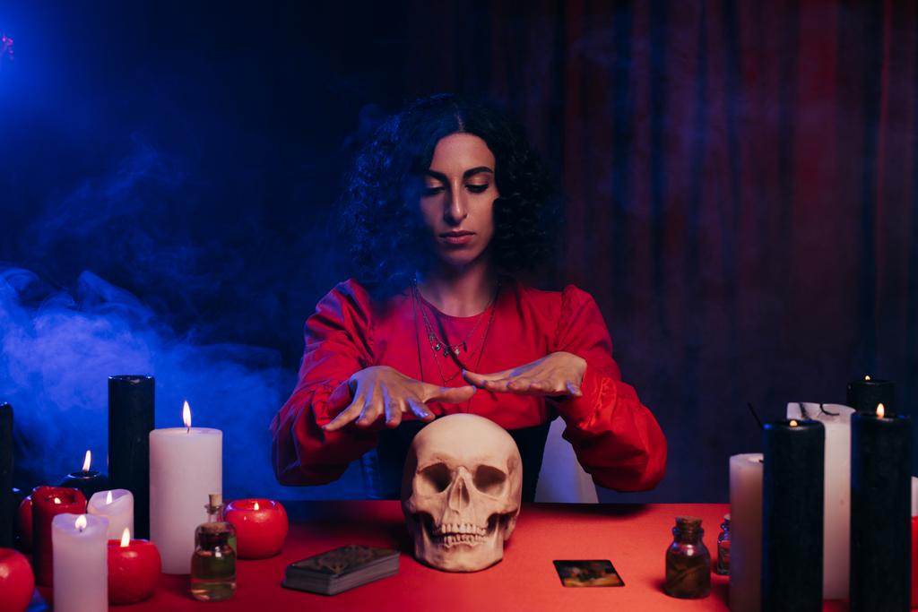 brunette oracle during magic session near skull and burning candles on dark background with blue smoke - Zdjęcie, obraz