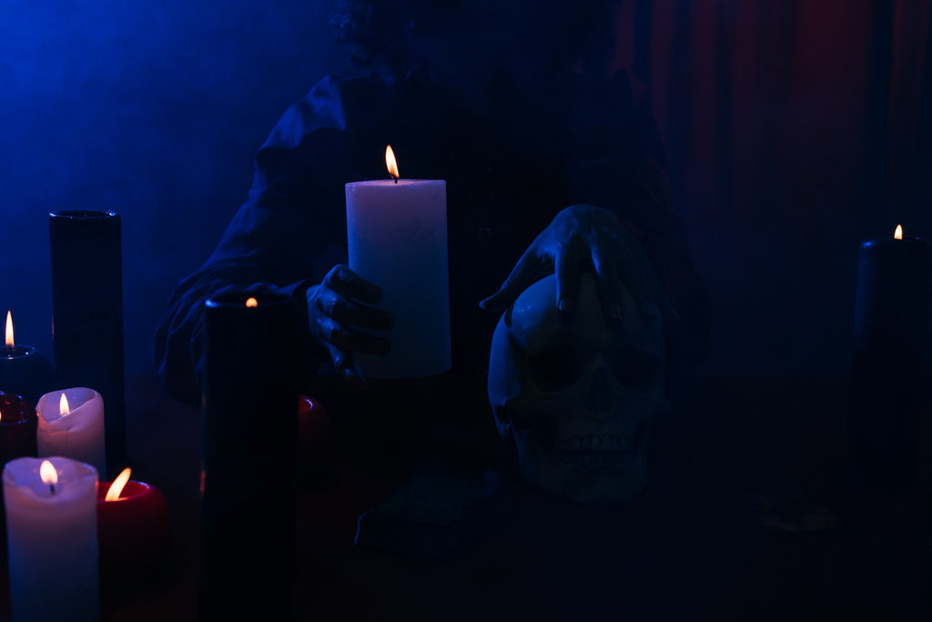 cropped view of oracle with burning candle and skull during magic session in darkness near blue smoke - Foto, Imagen