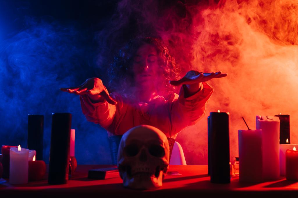 oracle with closed eyes holding hands above skull during spiritual session in darkness near colorful smoke - Valokuva, kuva