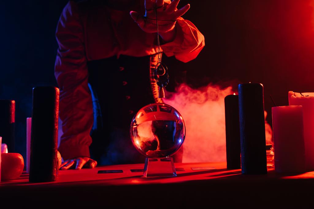 cropped view of medium holding amulet over crystal ball on black background with smoke - 写真・画像