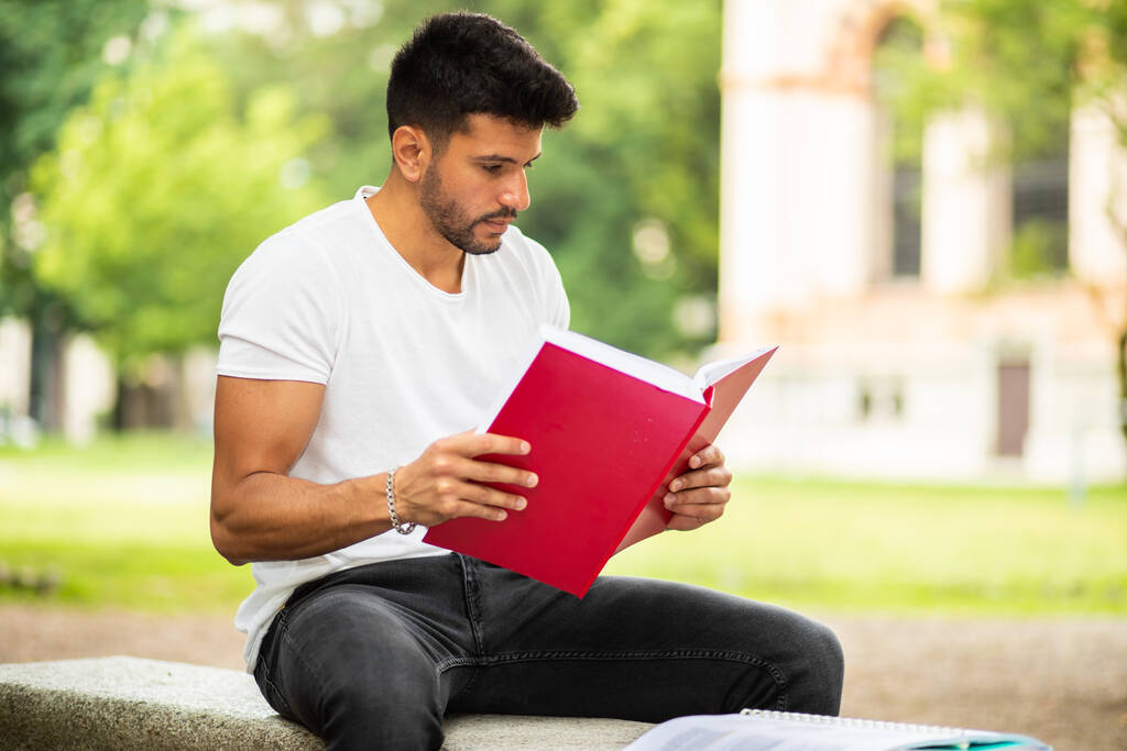 Komea nuori mies lukemassa kirjaa penkillä puistossa
 - Valokuva, kuva
