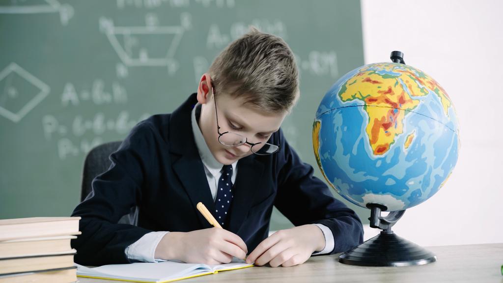 schoolboy in glasses writing in notebook near globe and books on desk - Foto, Bild