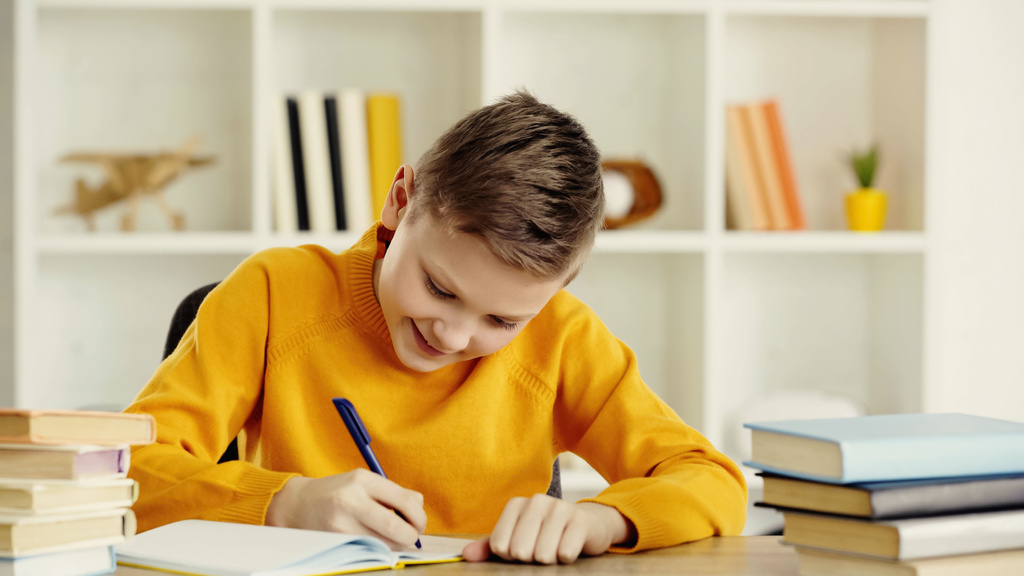 smiling preteen boy writing in notebook while doing homework near books on wooden table at home - Foto, Bild