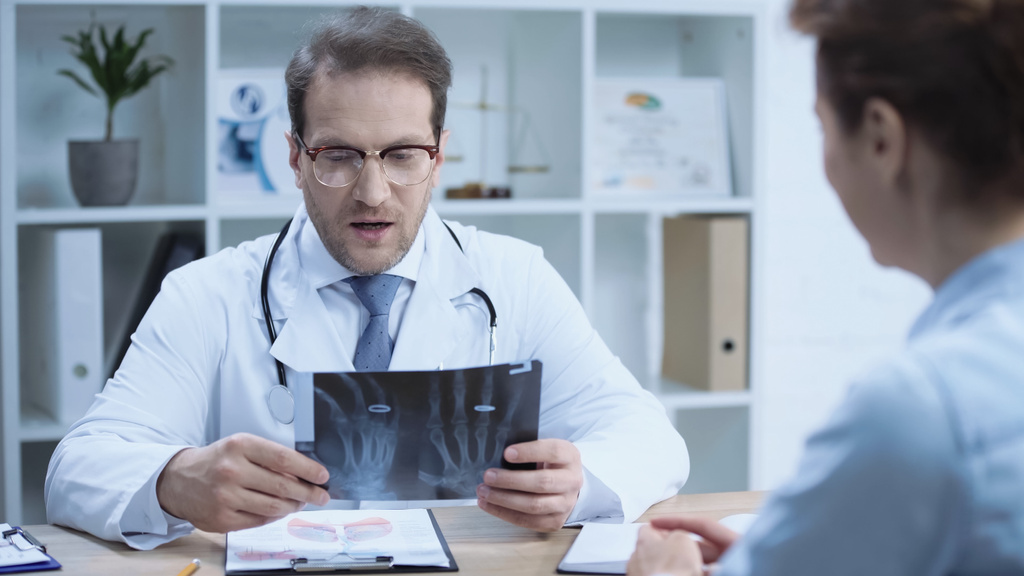 serious doctor sitting at workplace while looking at x-ray scan near woman on blurred foreground - Foto, Imagen