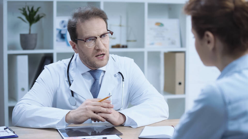 serious doctor in glasses and white coat sitting with clenched hands while talking with patient on blurred foreground  - 写真・画像