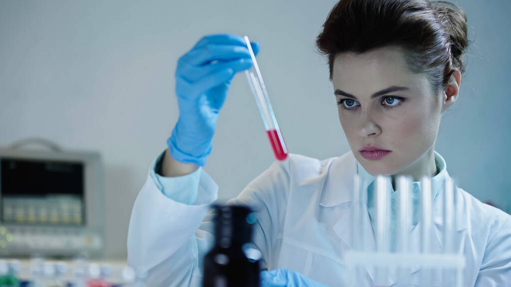 pretty scientist looking at red liquid in test tube near blurred medical equipment  - Foto, Imagem