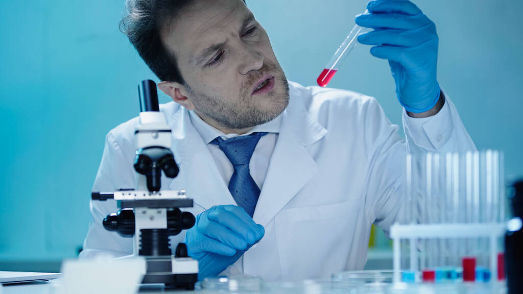 concentrated scientist looking at test tube with red substance near microscope in laboratory  - Foto, Imagen