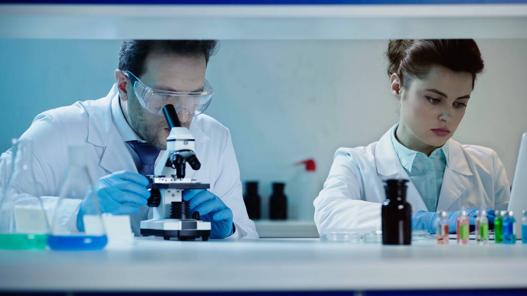 scientist in goggles looking through microscope near colleague using laptop in laboratory - Φωτογραφία, εικόνα