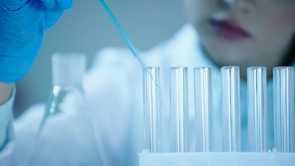 partial view of blurred scientist adding blue liquid into clean test tube in laboratory  - Foto, afbeelding