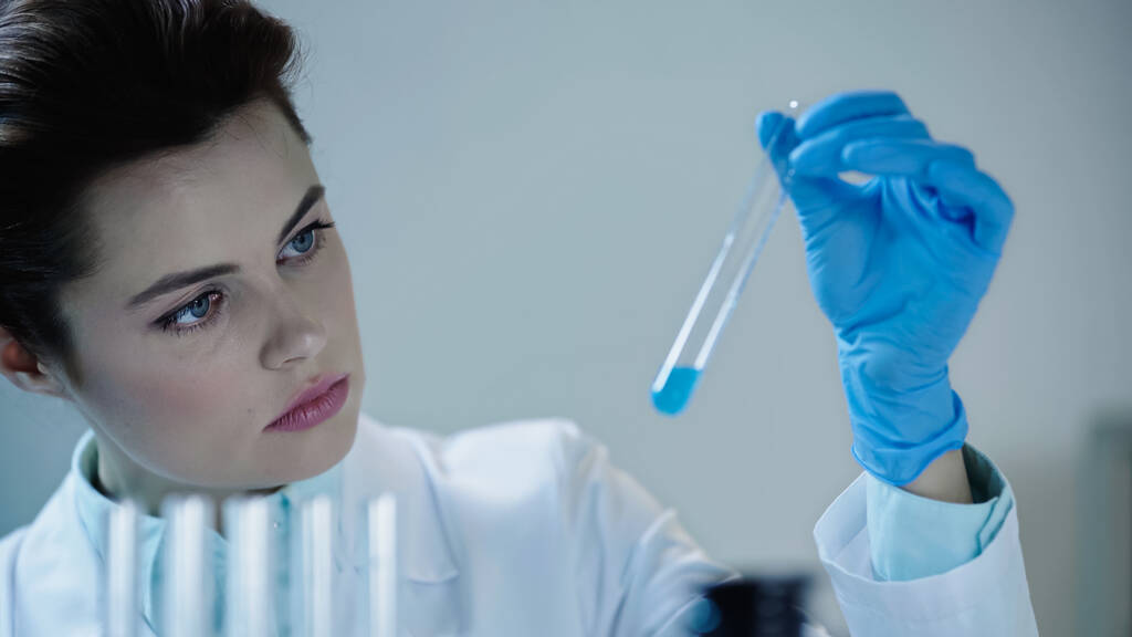 attentive female scientist looking at blue liquid in test tube and writing down results  - Foto, Bild