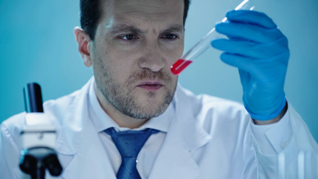 male scientist looking at test tube with red liquid in clinical laboratory - Photo, image