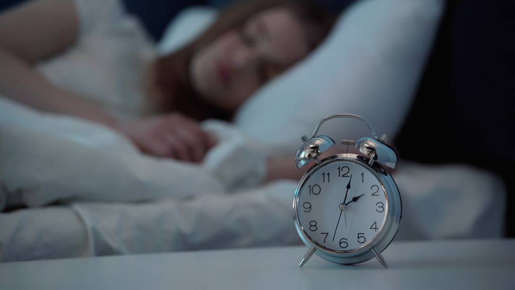Alarm clock on bedside table near blurred woman sleeping on bed  - Foto, Imagen