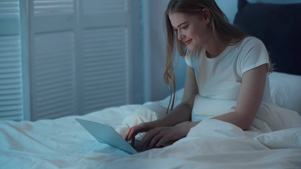 Весела жінка використовує ноутбук, сидячи на ліжку вночі
  - Фото, зображення