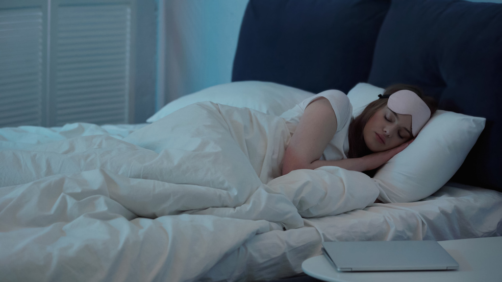Woman in mask sleeping on bed near laptop on bedside table  - Fotó, kép
