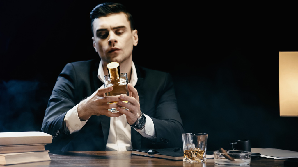 Businessman holding bottle of whiskey near books and cigar on table on black  - Fotografie, Obrázek