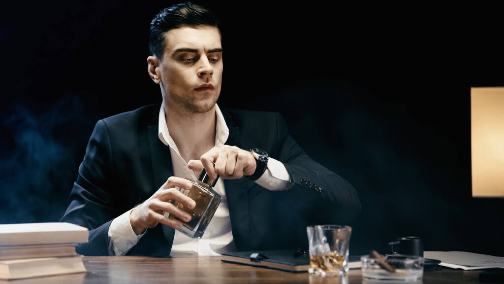 Businessman holding bottle of whiskey near books and ashtray on table on black  - Photo, image
