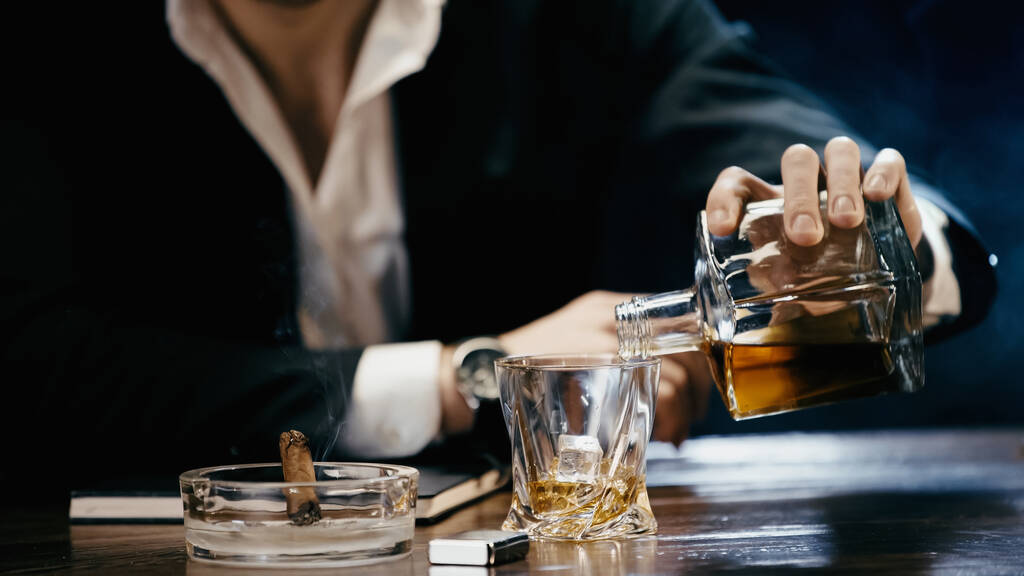 Cropped view of businessman pouring whiskey near cigar and lighter on black  - Foto, imagen
