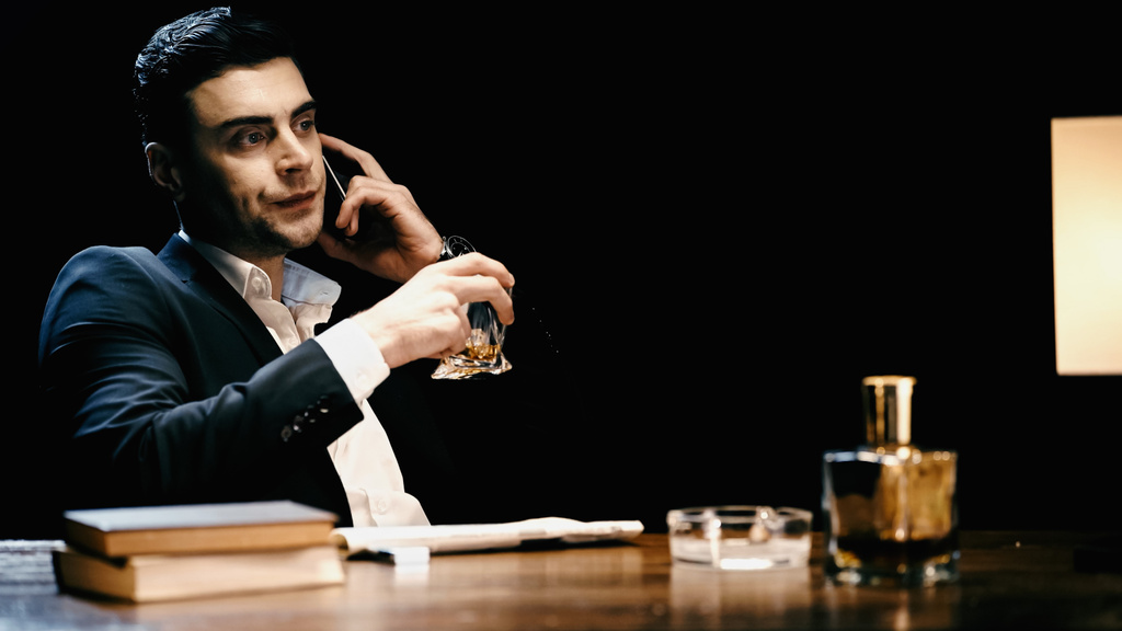 незадоволений бізнесмен тримає віскі і розмовляє на мобільному телефоні біля книг і пляшки ізольовані на чорному
  - Фото, зображення