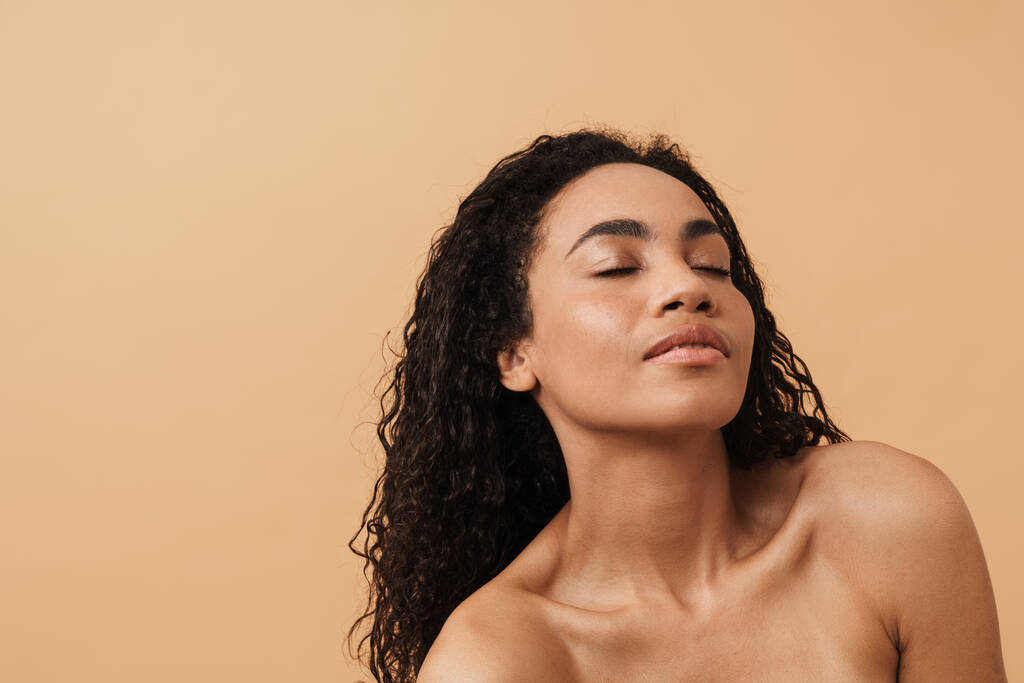 Shirtless black woman with wavy hair posing on camera isolated over beige background - Fotografie, Obrázek