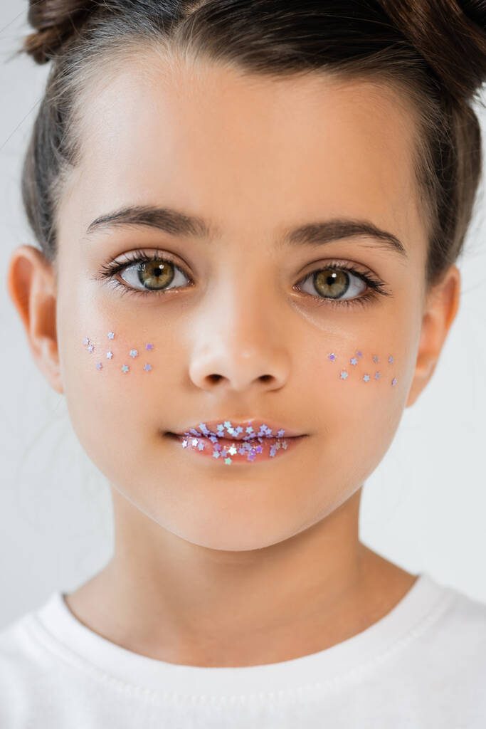 portrait of adorable girl with sparkling glitter stars on face and lips looking at camera isolated on grey - 写真・画像