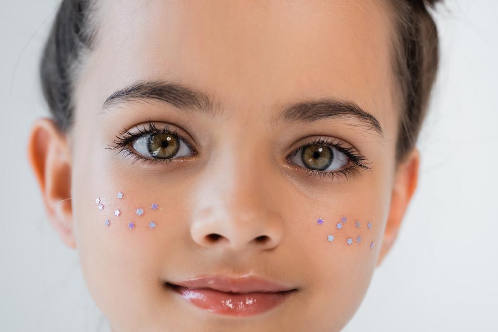portrait of smiling girl with hazel eyes and sparkling glitter stars on cheeks looking at camera isolated on grey - Foto, immagini