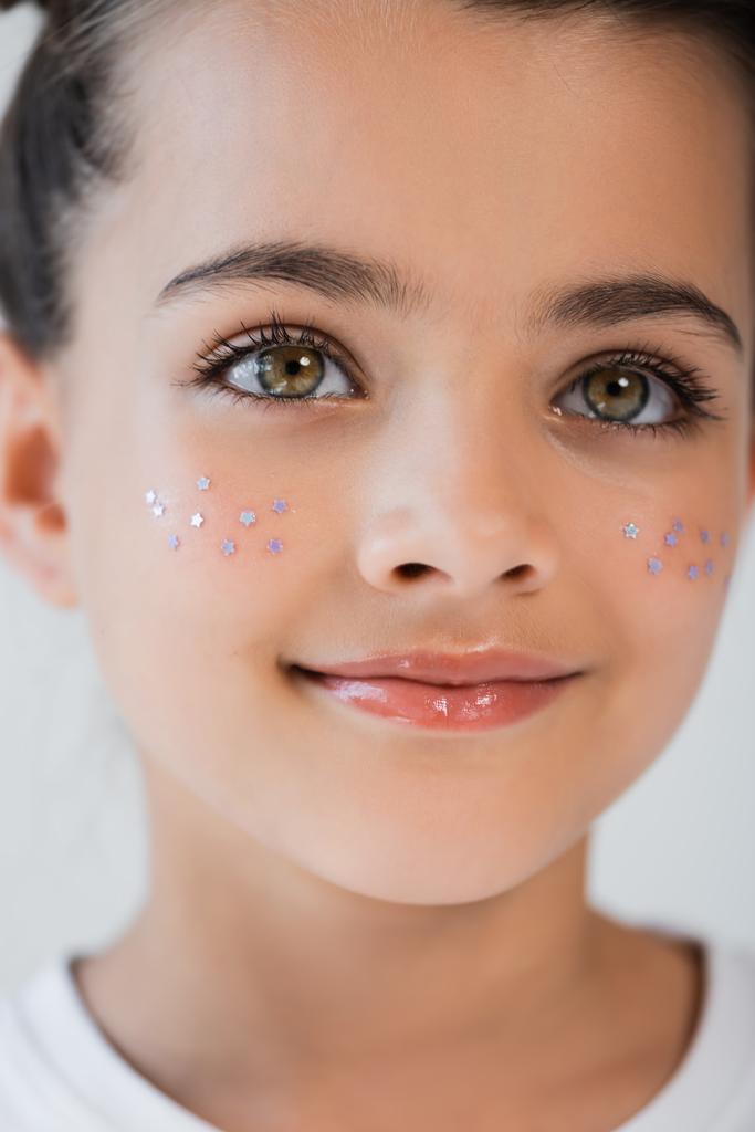 close up view of cheerful girl with hazel eyes and sparkling glitter stars on cheeks isolated on grey - Фото, изображение