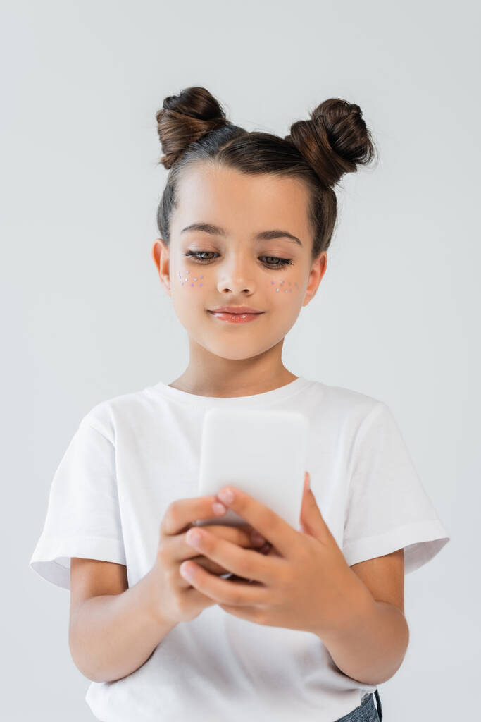 cheerful girl with glitter stars on cheeks using smartphone isolated on grey - Fotó, kép