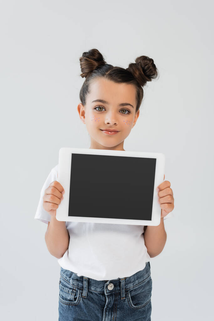 adorable girl with glitter stars on cheeks smiling while holding digital tablet with blank screen isolated on grey - Фото, изображение