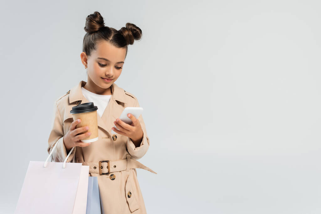 cute girl in trench coat holding paper cup and shopping bags while using smartphone isolated on grey  - Foto, afbeelding
