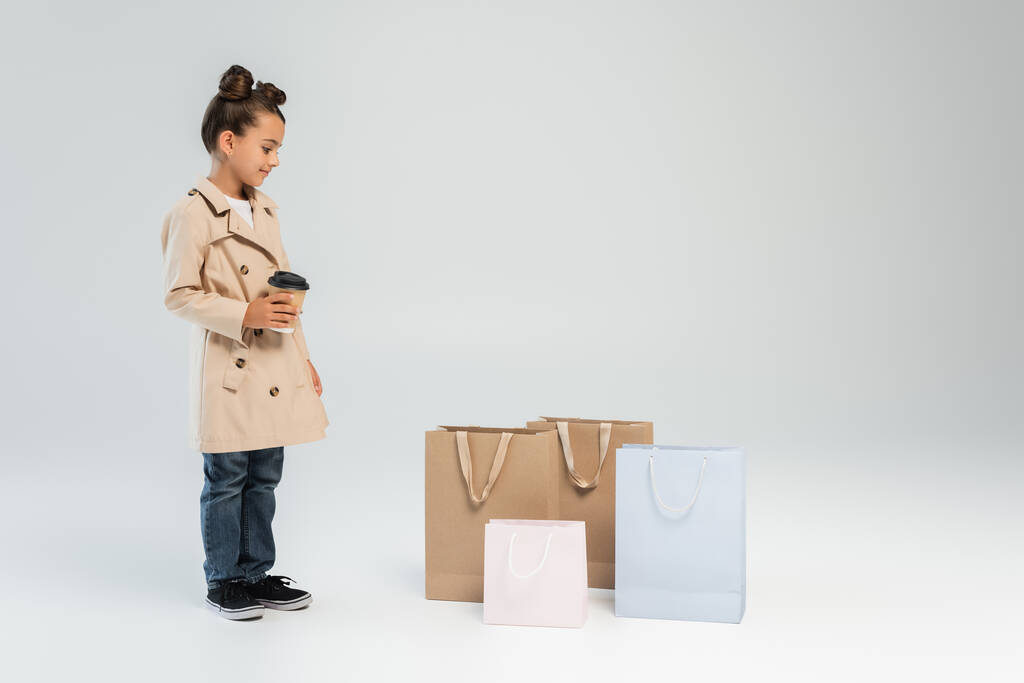 happy girl in trench coat and jeans holding paper cup and looking at shopping bags on grey  - Foto, Bild