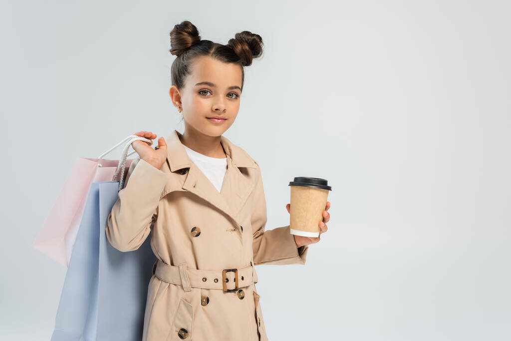 adorable girl in trench coat holding takeaway drink and shopping bags isolated on grey  - Photo, Image