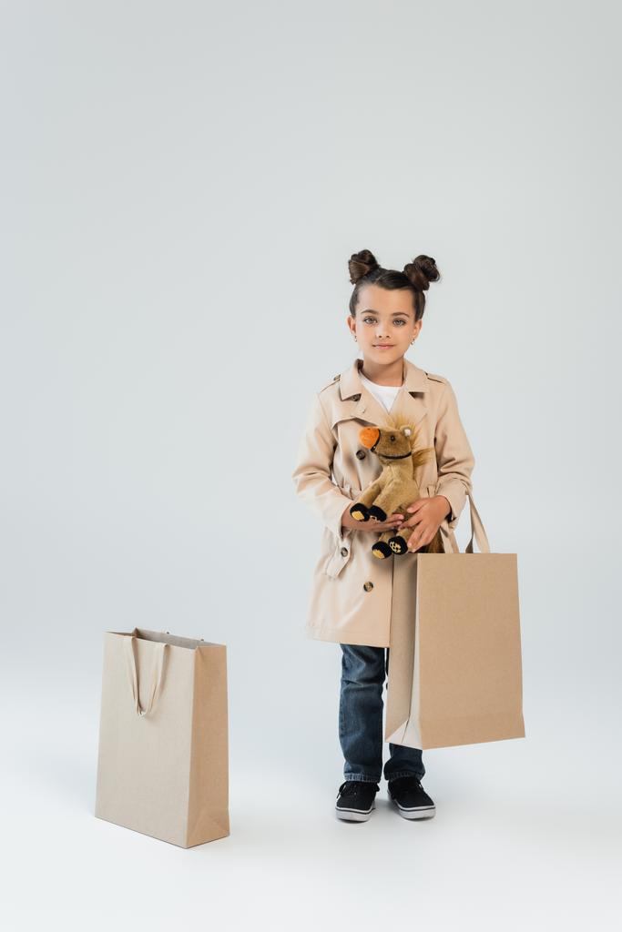 full length of cheerful kid in trench coat and jeans holding soft toy and shopping bag on grey - Foto, afbeelding