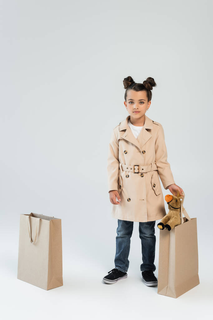 full length of stylish kid in trench coat and jeans holding shopping bag with toy horse on grey - Photo, image