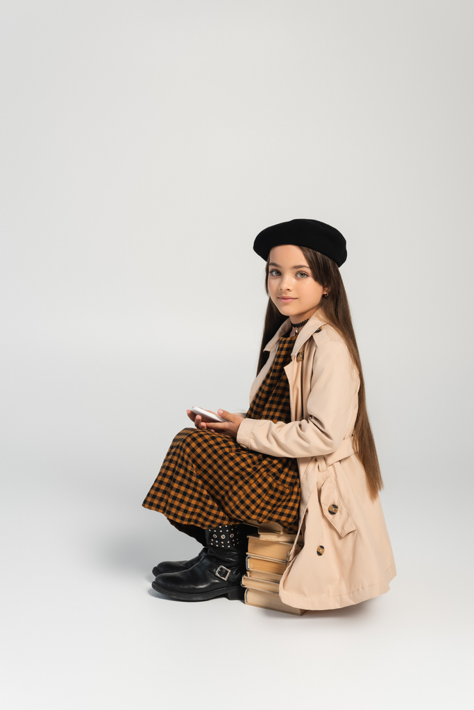 full length of cute kid in stylish autumnal outfit and beret sitting on stacked books and holding smartphone on grey - Foto, afbeelding