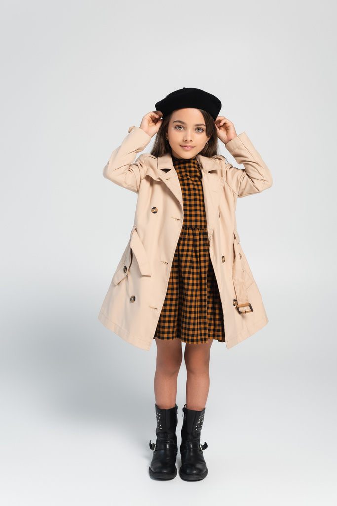 full length of cheerful girl in stylish trench coat adjusting beret while standing on grey - Foto, Imagen