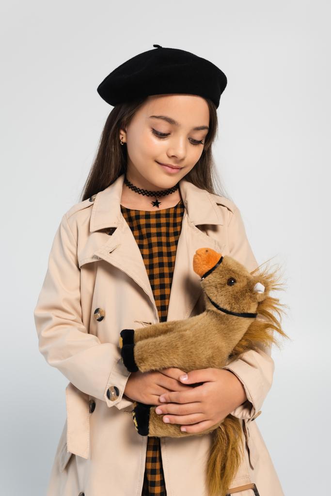cheerful girl in stylish trench coat and beret standing with toy horse isolated on grey - Фото, изображение