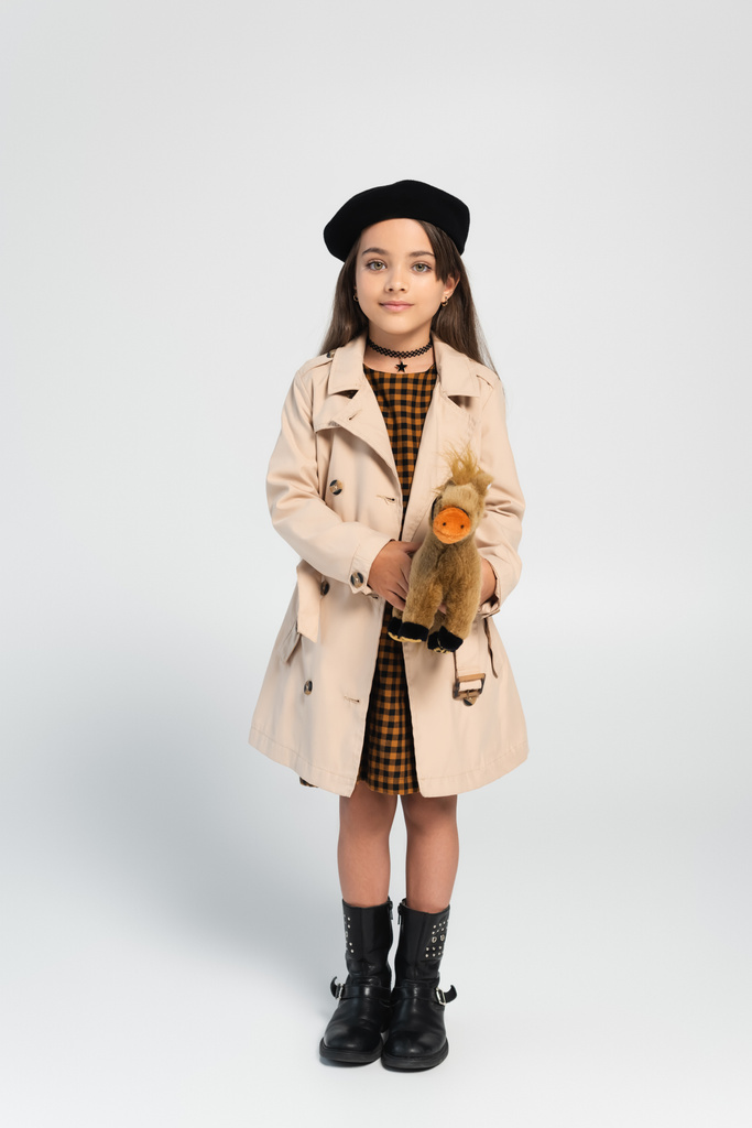 full length of cute girl in stylish trench coat and beret standing with toy horse on grey - Foto, Imagen