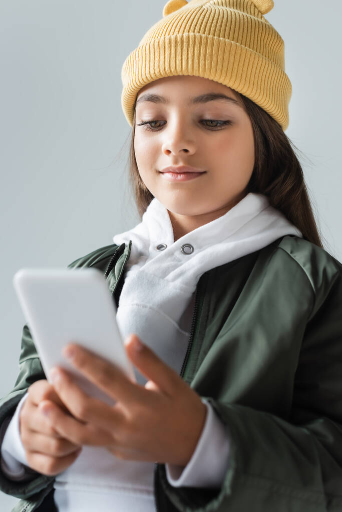 cheerful kid in trendy autumnal outfit and beanie hat using smartphone isolated on grey - Foto, afbeelding