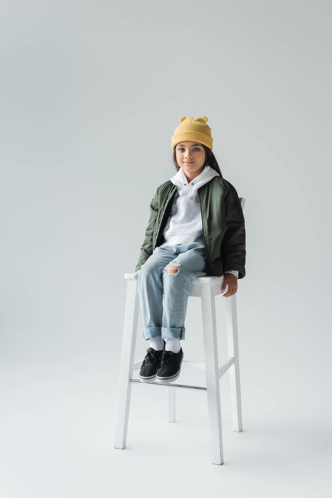 full length of cute girl in trendy autumnal outfit sitting on high chair on grey - Zdjęcie, obraz
