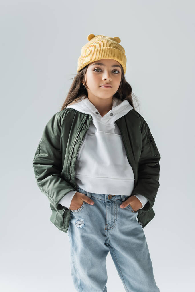 portrait of cute kid in yellow beanie hat and stylish autumnal outfit posing with hands in pockets isolated on grey  - Photo, image