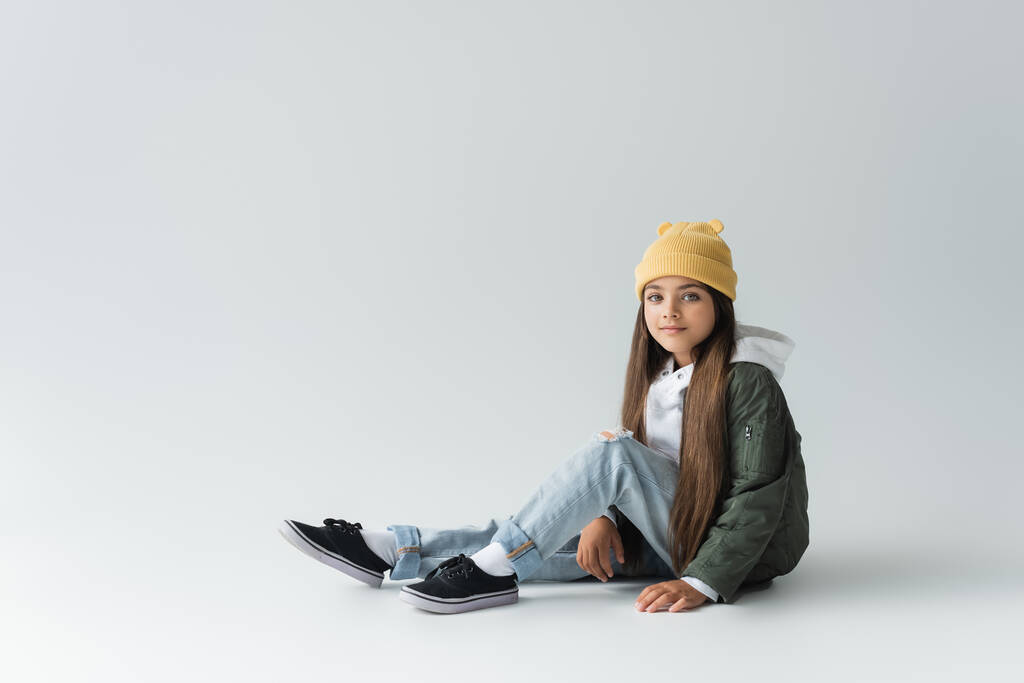 full length of adorable girl in trendy autumnal outfit and beanie hat sitting on grey - 写真・画像